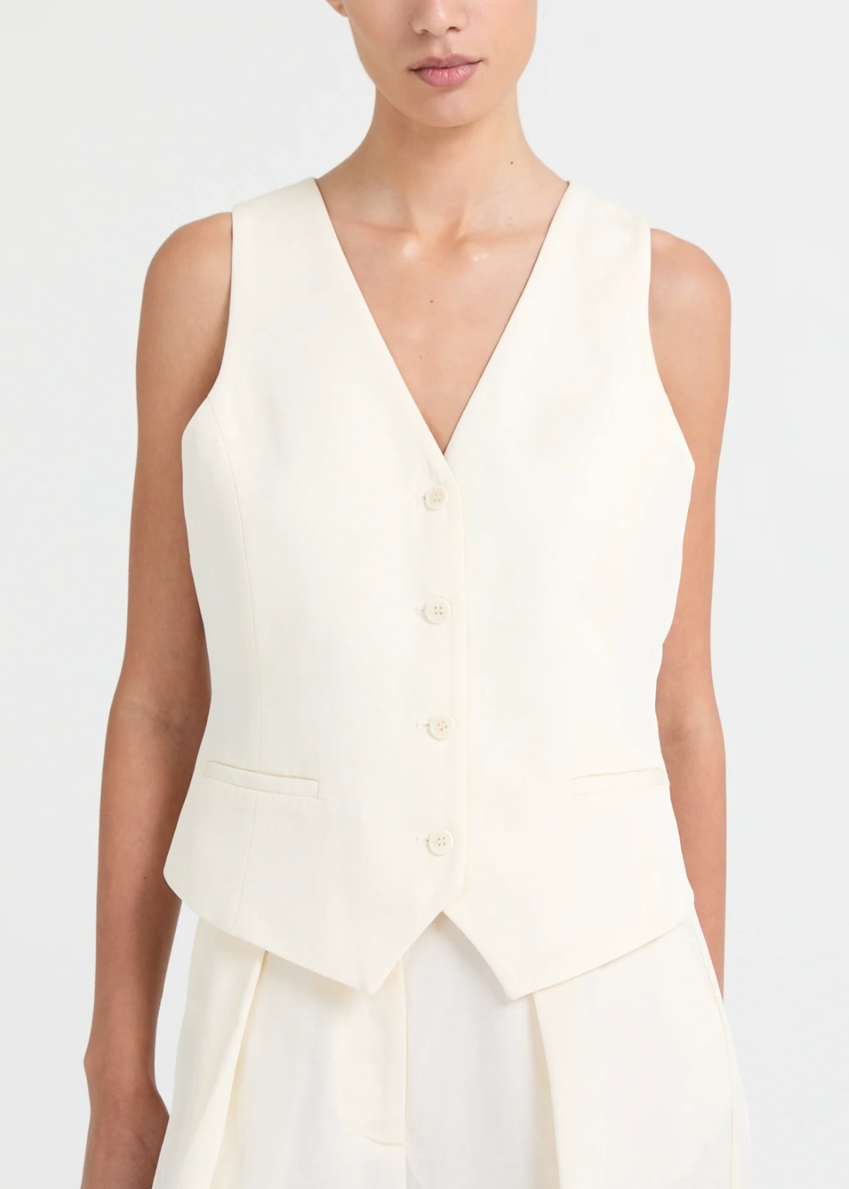 A tailored waistcoat cut in a softly structured, fluid fabric. Designed with a clean V-neckline and a slim, elongated silhouette, it features tonal buttons and subtle welt pockets that enhance its refined construction. The pointed hem adds definition, creating a sharp yet feminine line. A modern layering piece that balances tailoring with ease. Wear it on its own or styled with the matching suit for a sophisticated look.